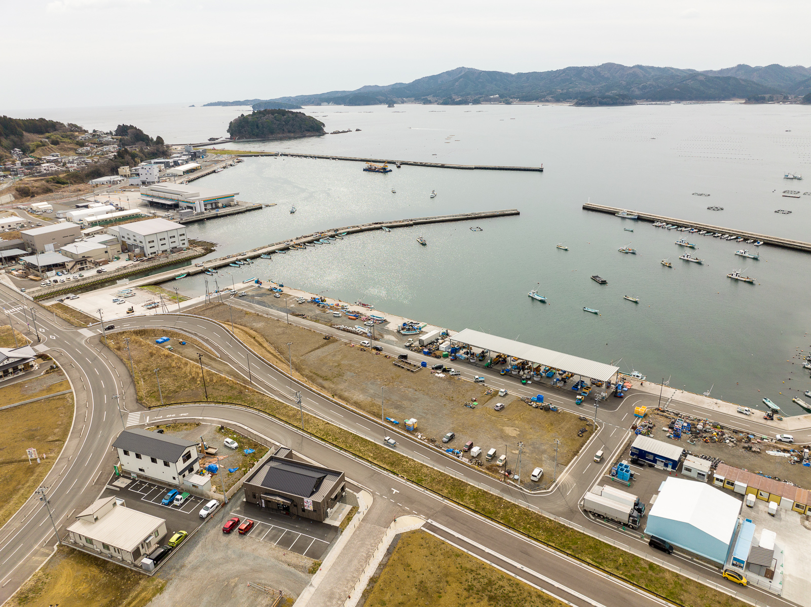 右側の海沿いには漁業施設や倉庫が建ち並び、多くの小型漁船が岸に停泊しており、湾内は波が穏やかで、背景は山と海の境界が広がる漁港と作業施設の昼間の風景を高台から定点カメラで撮影した写真
