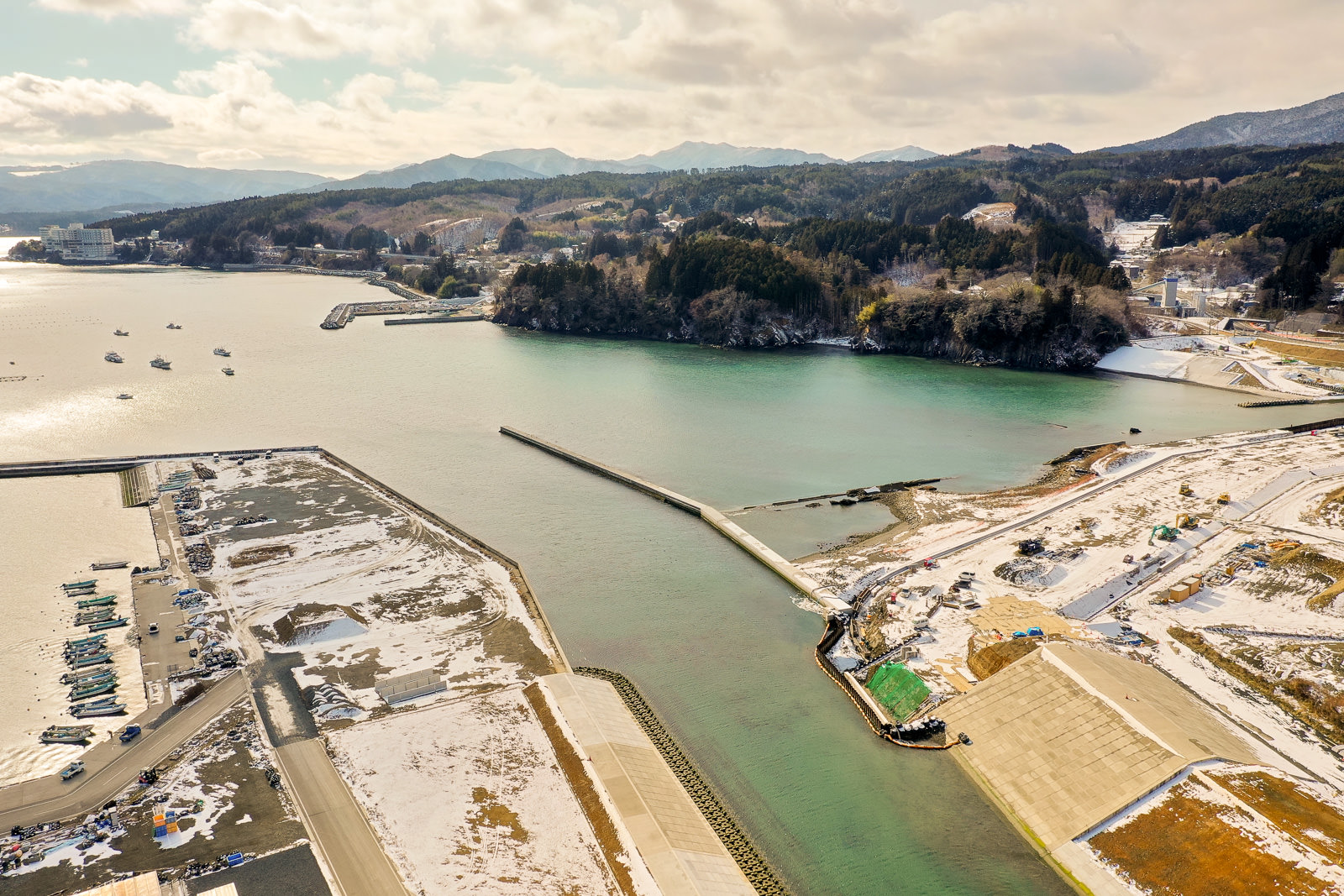 港を中心に雪が積もった陸地が広がり、複数の船が桟橋に停泊しており、防波堤が海に向かって伸びて波を防いでいる様子が見え、周囲には森林に覆われた丘や山々が連なり、海岸沿いにはいくつかの建物が点在している冬の海辺の港の写真