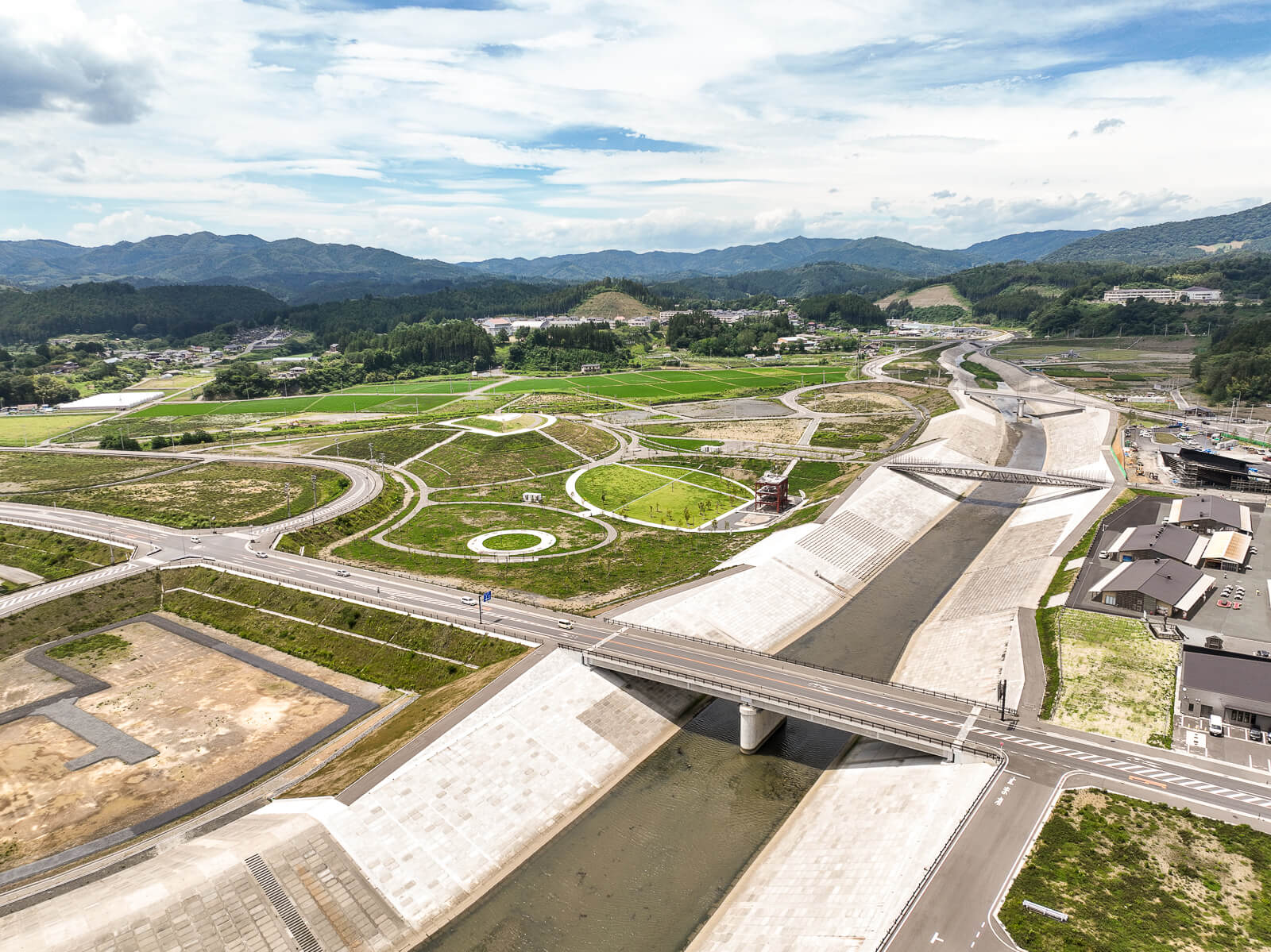 護岸工事された川沿いに、整備された舗装道路と緑地、遠くに山並みが広がる開発途中の地域の昼間の風景を高台から定点カメラで撮影した写真