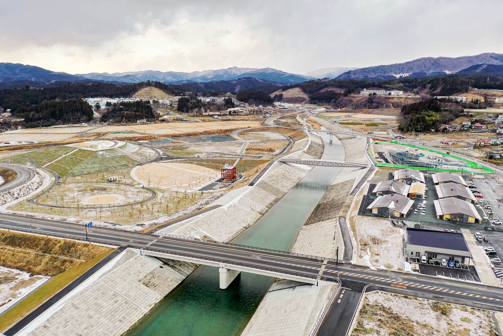 中央に流れる川に沿って整備された河川敷と橋、左側には丸い広場や植栽が見られ、右側には駐車場と新しい建物群が並んでいる冬の昼間の風景を高台から定点カメラで撮影した写真