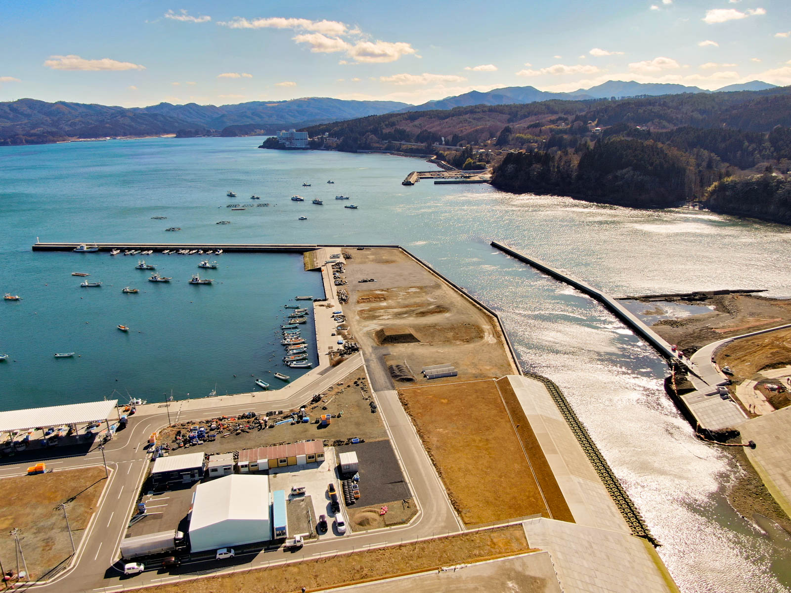 港を中心とした海岸地域を上空から見下ろした風景が広がっており、水面には複数の船が停泊し、防波堤に囲まれた港があり、陸地には建物や道路、広場が点在し、背景には山々が広がり、空には雲が部分的に浮かぶ写真