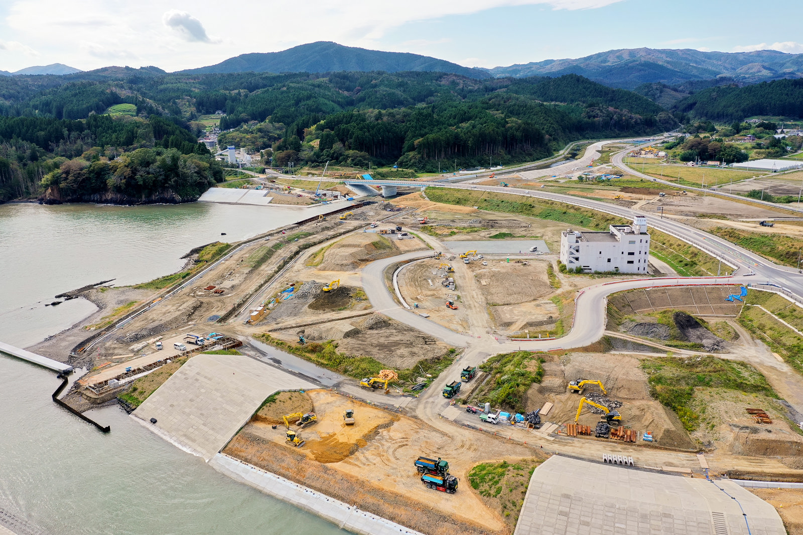 水辺に隣接する工事現場を空から捉えた風景で、複数の建設車両や機材が点在し、整地された区域と土の道路が広がり、背景には緑の丘や山々、遠くに建物やインフラが見える開発中の地域の写真