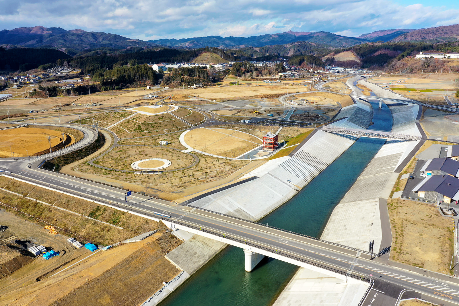 コンクリートで護岸された川が流れる風景を上空から撮影したもので、川には橋が架かり、その周囲には道路や地形に描かれた円形の模様が見られ、背景には丘や山が広がり、建物も点在し、空には雲が浮かぶ川沿い地域の写真
