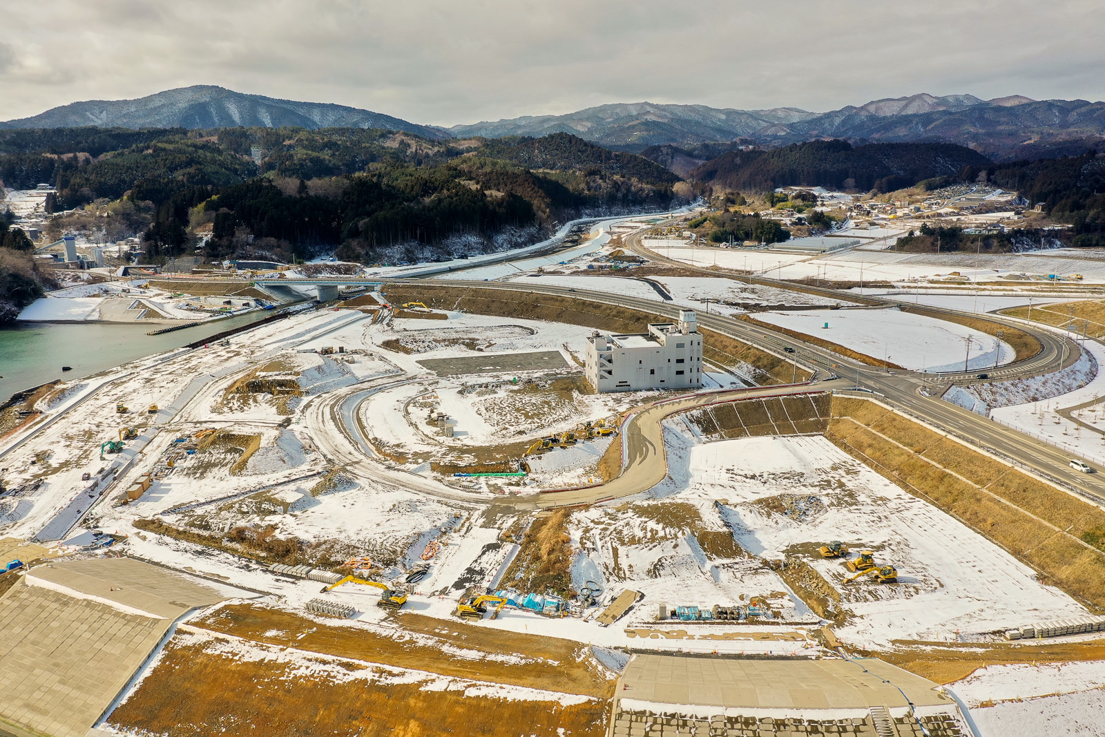 雪が積もった風景の中に曲がりくねった道路と川が流れ、橋が架かっており、複数の建設車両や重機が点在し、中央付近には大きな建物があり、背景には木々に覆われた山々が広がっている冬の工事現場周辺の写真