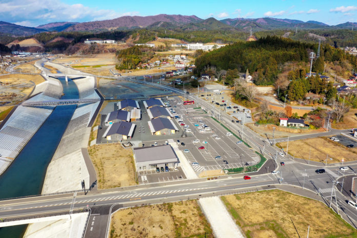 川沿いに広がる広い駐車場の奥に、複数の建物が並んでおり、周囲には緑が点在している風景の写真