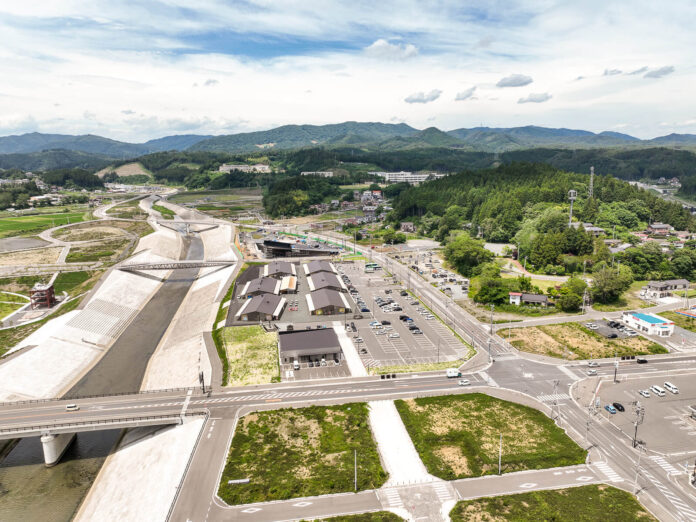 広い川の護岸工事が施された川沿いに整備された大きな駐車場と複数の建物が立ち並び、その奥に山々と町並みが広がっている昼間の風景を高台から定点カメラで撮影した写真
