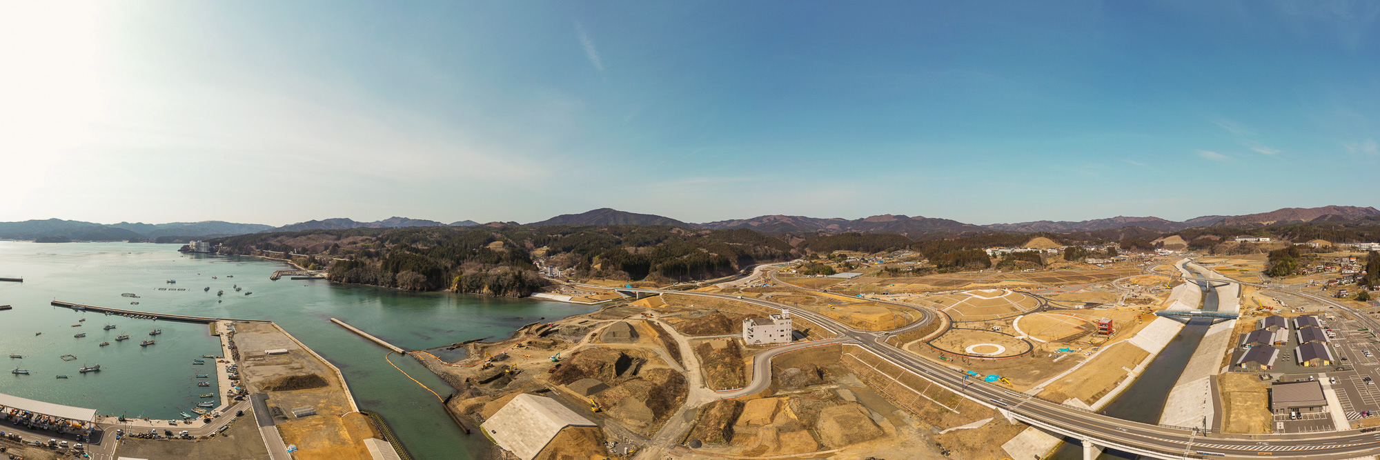 左側に船が停泊する海が広がり、その隣接地では複数の区画が整地され、道路や建物などのインフラが整備されつつある開発中の沿岸地域を広く見渡せる、背景に木々に覆われた山々が広がる晴天の海辺の開発地の写真