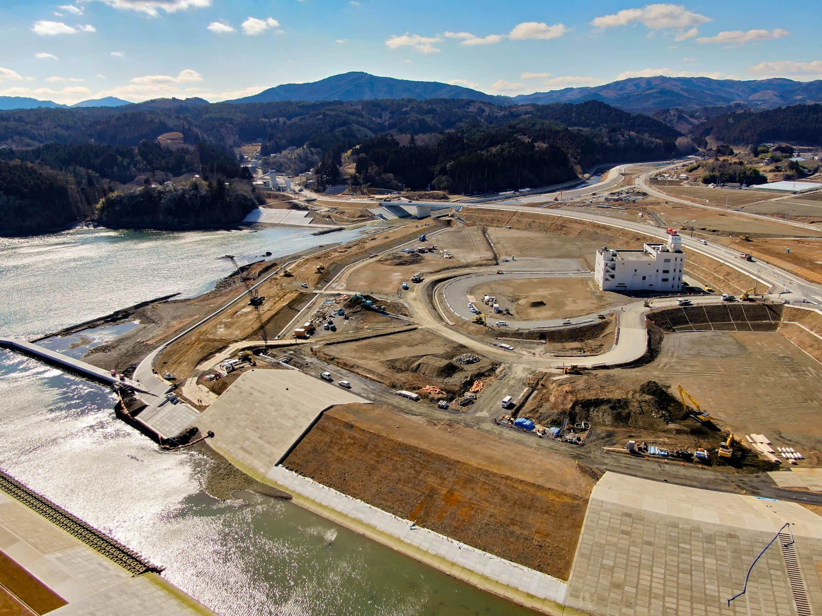 建設現場を上空から見下ろした風景が広がっており、複数の道路と構造物、車両がある大きな円形の区画、白い多層建物が見られ、背景には山々が広がり、空にはいくつかの雲が浮かぶ晴天の写真