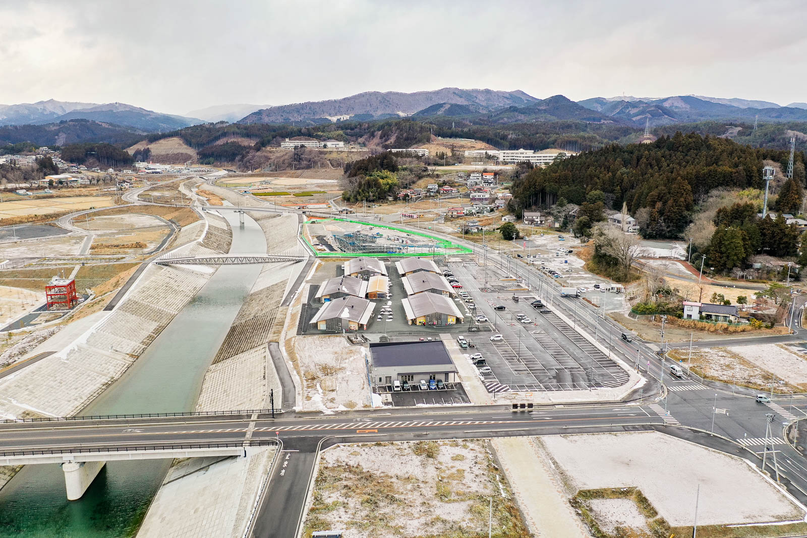 川沿いに整備された道路と複数の新築建物、さらにその奥には民家や山が見え、手前には交差点と駐車場が広がっている昼間の風景を高台から定点カメラで撮影した写真