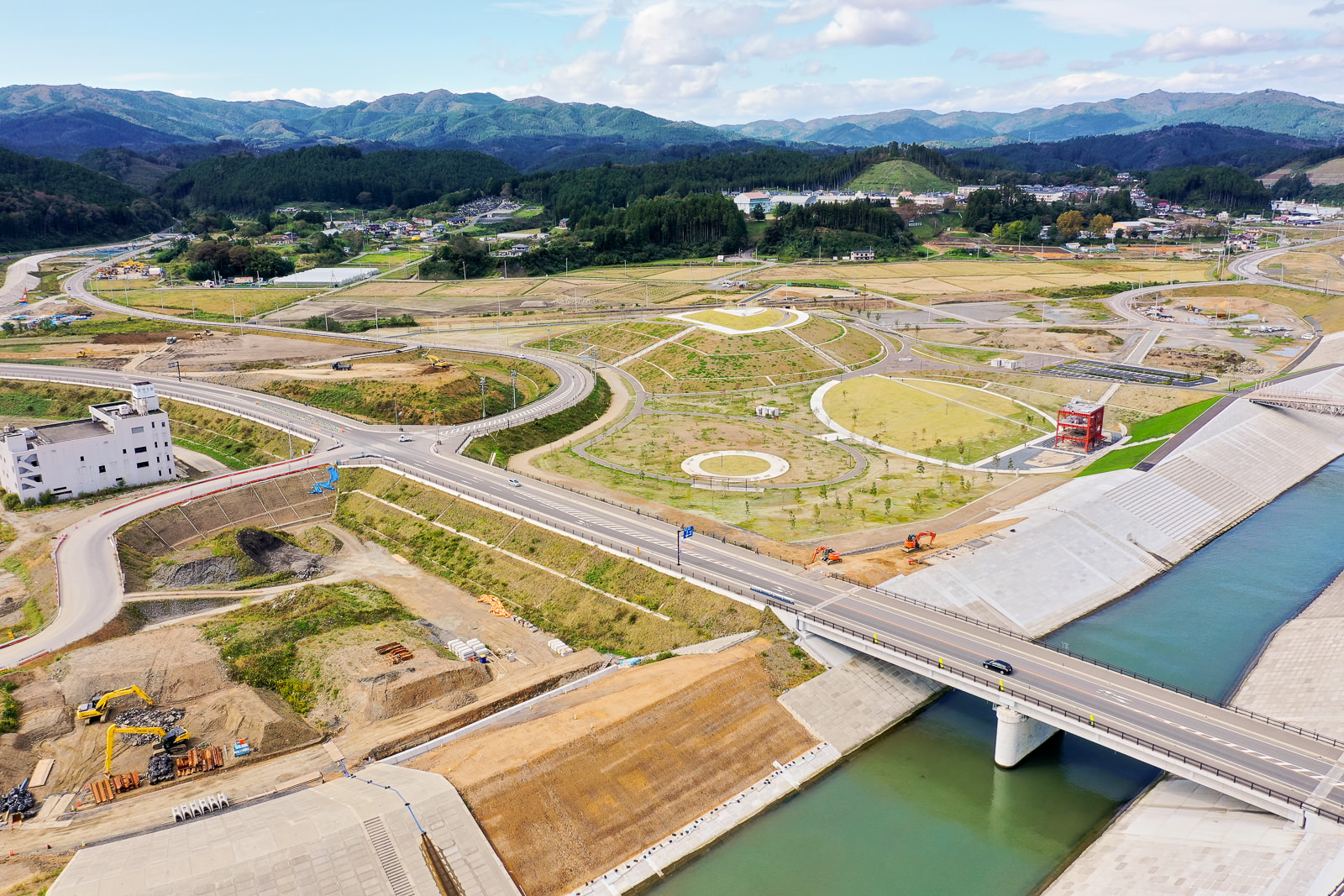 川に架かる橋を中心に、周囲に道路や歩道が曲がりくねって広がり、橋の近くには建設車両や機材が点在し、背景には丘や山々、点在する建物と緑が広がる開発中の橋周辺地域の写真