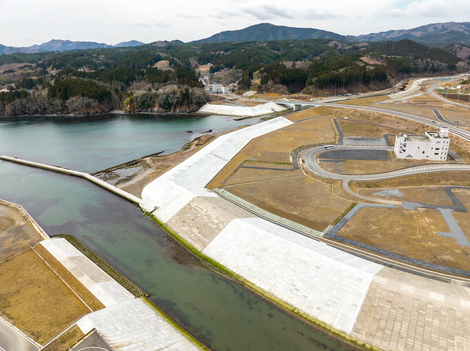 手前に広い川と白く整備された護岸が見え、奥には少し曲がった防波堤と海がつながっており、右手には新しく舗装された道路と白い建物があり、背景には山が重なる川の河口付近と防波堤の昼間の風景を高台から定点カメラで撮影した写真