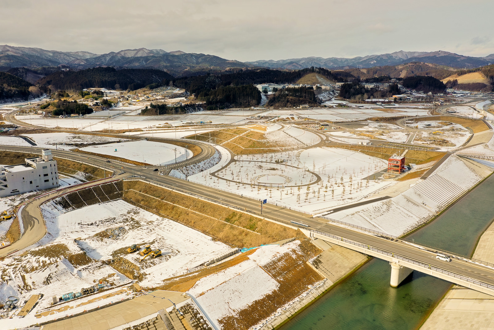 雪が積もった風景の中に川が流れ、その上に橋が架かっており、周囲には道路や小道が広がり、建物が点在し、背景には雪に覆われた山や丘が連なっている冬の山間の町の写真