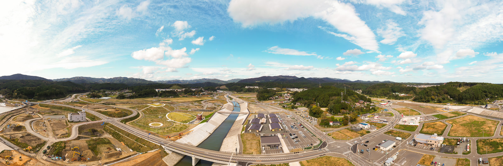 開発された地域と未開発の土地が混在する半都市的な風景を広く捉えた構図で、中央には川が流れ、複数の橋が横断し、周囲には道路や建物、空き地が広がり、背景には山々がそびえる風景写真