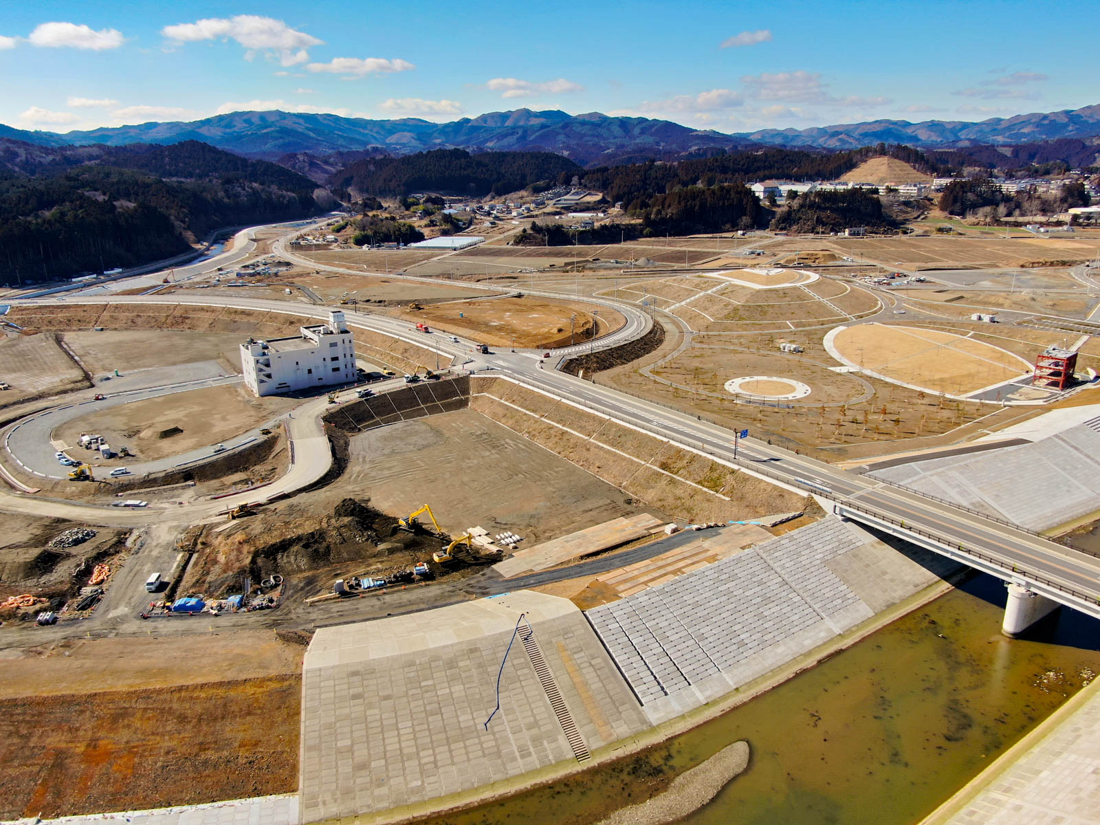 広大な建設現場が上空から写されており、中央付近には目立つ白い建物があり、その周囲には円形や長方形の区画が広がり、手前には水路に架かる橋があり、背景には山々が広がっていて、現場は開発初期段階で建設機材も見られる写真