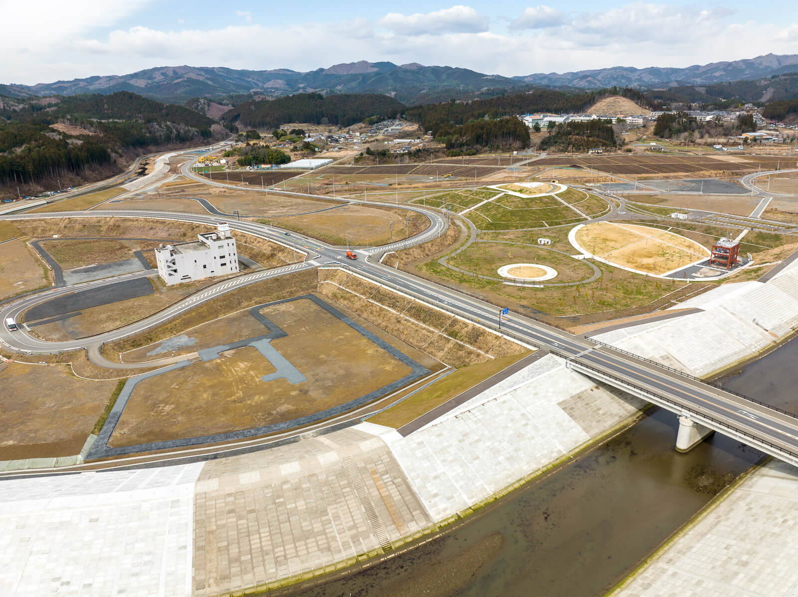 川を貫く道路の右側に緑地と円形の植栽がいくつかあり、左側には白い建物があり、背後には山並みが続く、全体が整然と整備された復興公園付近の昼間の風景を高台から定点カメラで撮影した写真