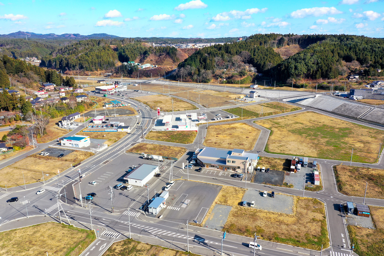 複数の道路が交差する農村地域を上空から撮影した風景で、ガソリンスタンドや小規模な商業施設、駐車場などの建物が点在し、周囲には広い畑や空き地が広がり、背景には森林に覆われた丘が見える農村地域の写真