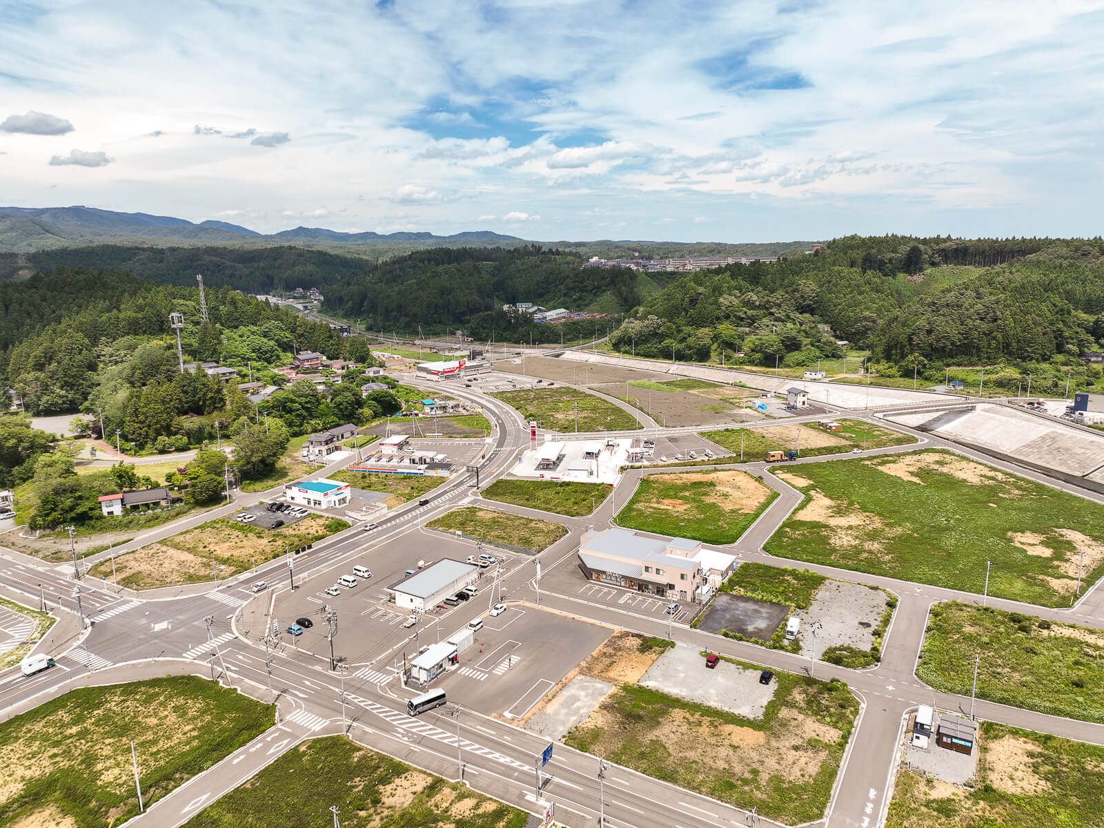 交差点の周囲に住宅や店舗が点在し、周囲を森林に囲まれ、整備された道路や空き地が広がっている町の昼間の風景を高台から定点カメラで撮影した写真