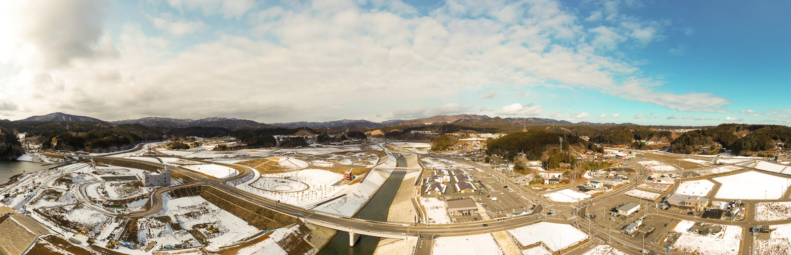 雪に覆われた田園地帯を広く見渡した風景で、中央には川が流れ、複数の橋がその上を横断しており、周囲には道路や建物、畑が広がり、背景には丘や山が連なり、空には雲が浮かぶ冬の農村地域の写真
