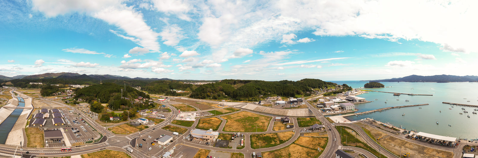 海に面した町を広く見渡す構図で、建物や道路、港が広がり、海には複数の船が浮かび、周囲には緑豊かな丘がある沿岸の町の写真