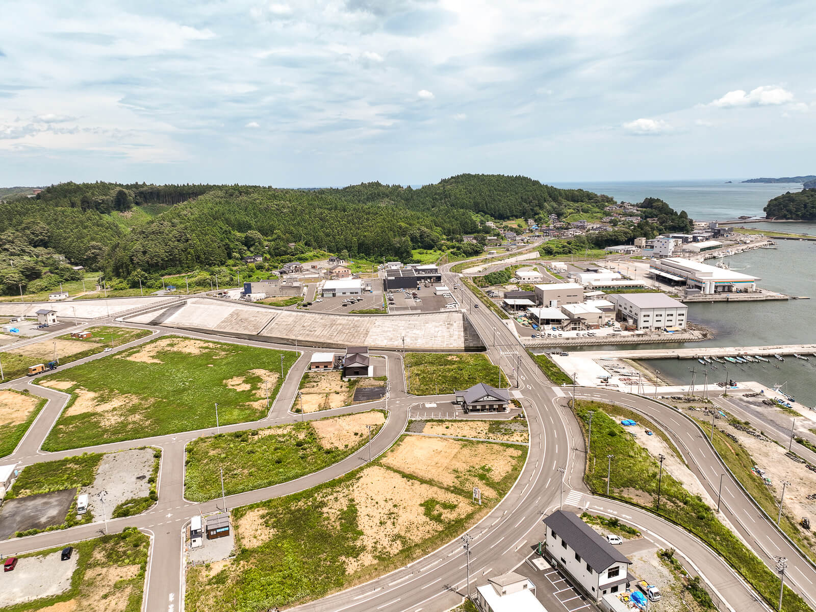 左下から港に向かって蛇行した道路が整備され、港に接した道路沿いに建物が並び、緑地や空き地が広がる地方都市の昼間の風景を高台から定点カメラで撮影した写真