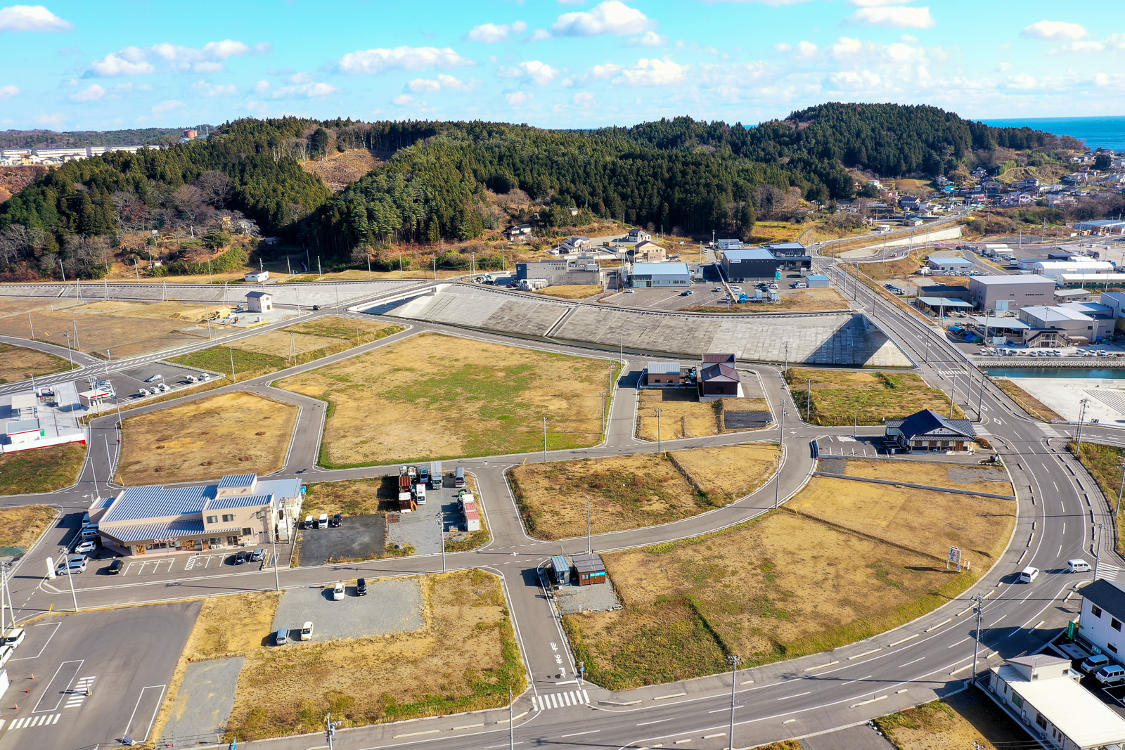 草地や舗装された区画が広がる地域を上空から撮影した風景で、複数の土地の区画や建物、道路が見られ、背景には木々に覆われた丘と建物が点在し、空には雲が浮かぶ写真