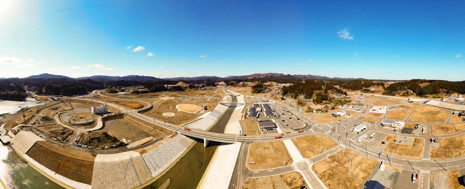 川や橋、道路、さまざまな建物が点在する広範囲の風景が上空から写されており、地域は開発中または最近整備された様子で、建設現場やインフラが見られ、空は雲がほとんどない晴天で、周囲には丘陵地とまばらな植生が広がっている写真