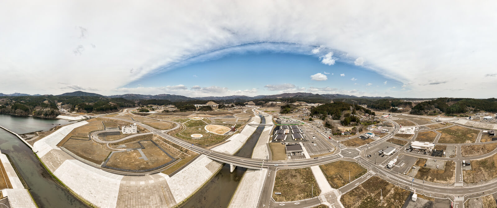 広範囲にわたる復興整備地域が写っており、中央には川と護岸がまっすぐ通っており、川の両側には道路と区画整理された空き地が広がり、奥には山々が連なる、復興工事が進められている町の全体の昼間の風景を高台から定点カメラで撮影した写真