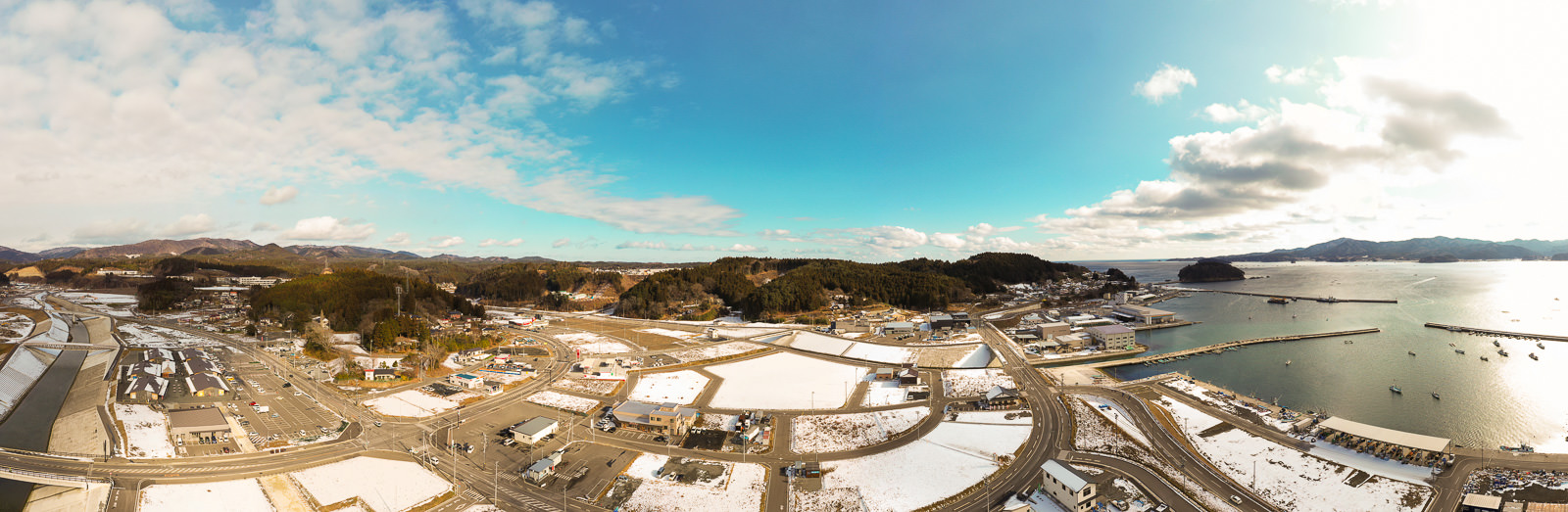 雪がまだらに積もった海沿いの町を広く見渡す構図で、複数の道路と建物が入り組む市街地が広がり、海には桟橋が伸び、そこに複数の船が停泊しており、背景には木々に覆われた丘や小さな山々が連なり、空は青く澄み渡りつつもところどころに雲が浮かんでいる写真