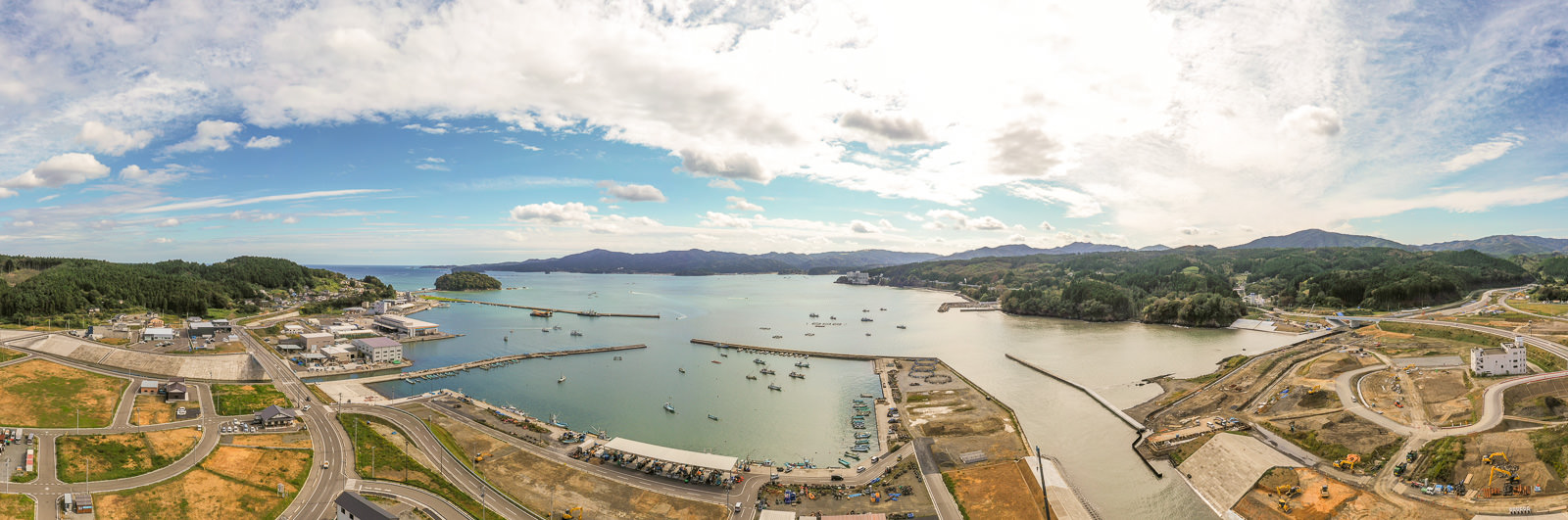 港を中心とした海沿いの地域を広く見渡す構図で、複数の船が停泊する海と、それを囲む道路や建物、緑豊かな丘が広がり、空には青空と雲が混在する沿岸地域の写真