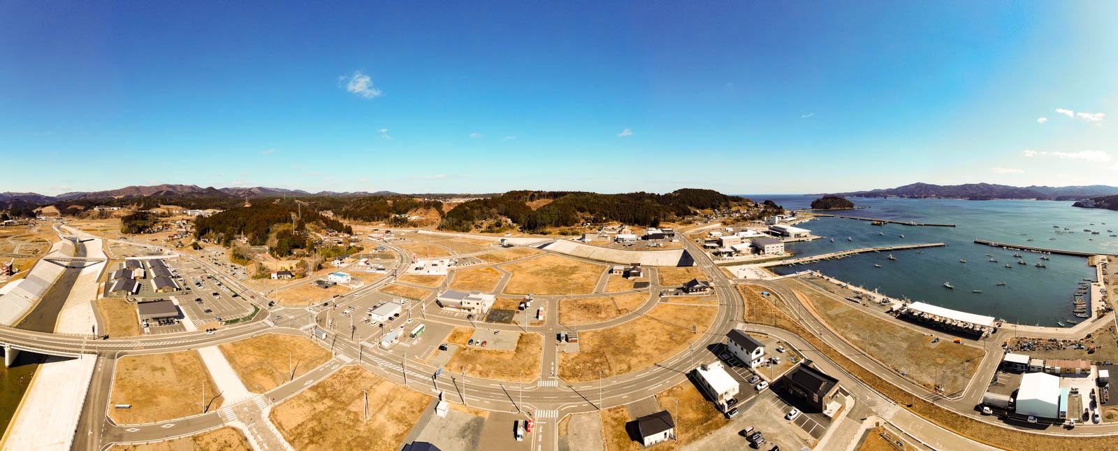 海沿いの町を上空から見下ろした風景が広がっており、複数の建物や道路、港があり、港にはいくつかの船が停泊していて、道路が交差する場所や広場、背景には緑地も見られ、空は雲がほとんどない晴天の写真