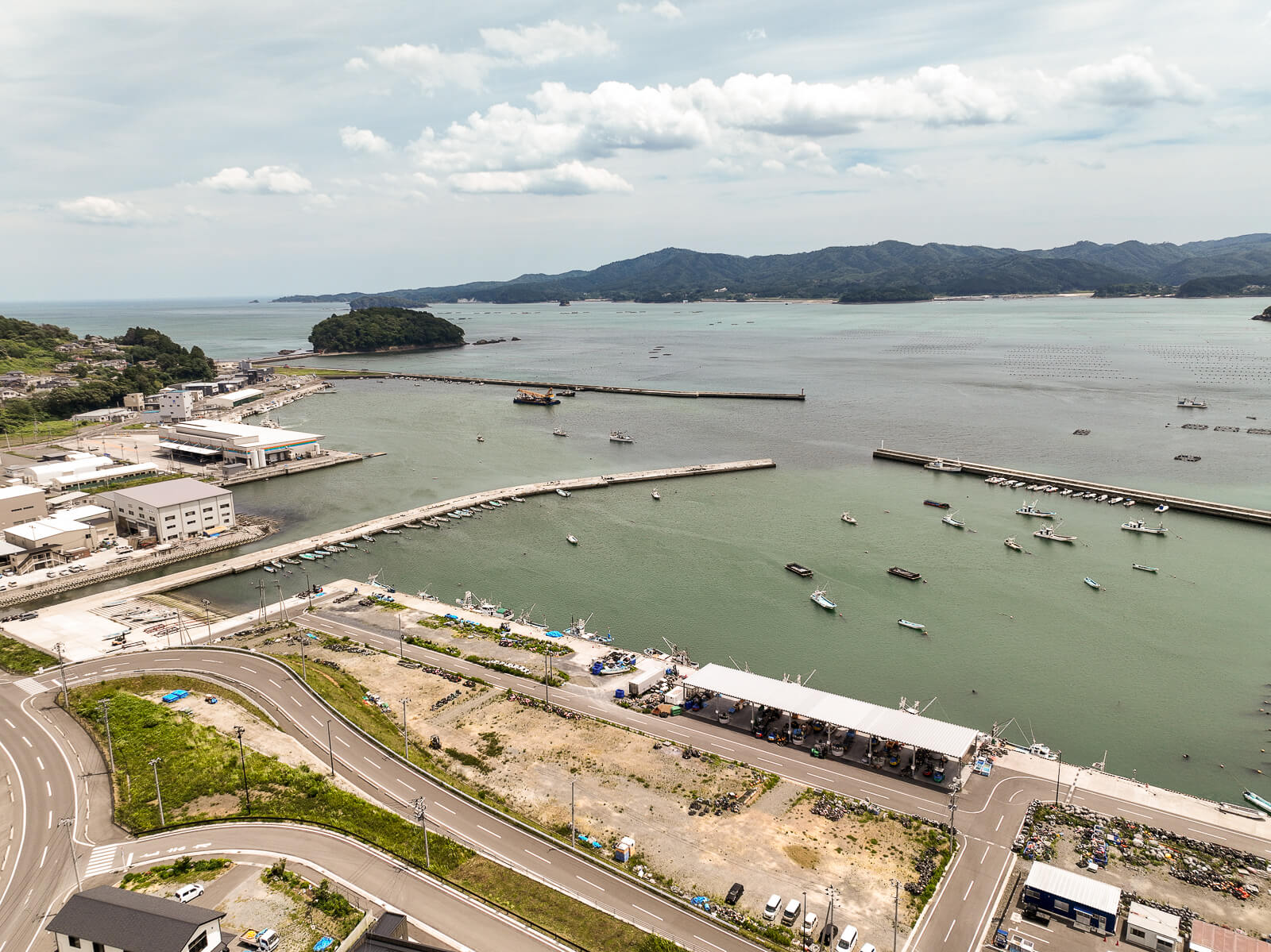 港湾の防波堤と停泊中の小型船舶、港に隣接する道路や建物、背景に山と海が広がる昼間の風景を高台から定点カメラで撮影した写真