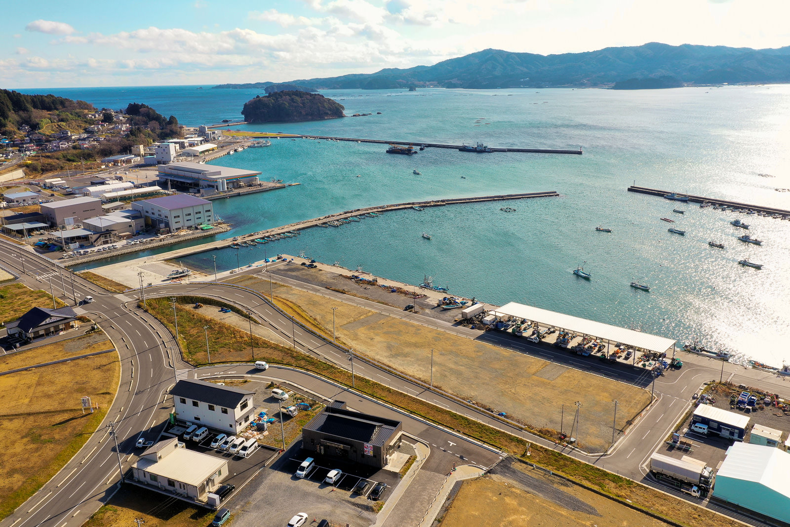 港を中心とした海沿いの町を上空から撮影した風景で、複数の桟橋が海に突き出し、船が係留されており、海岸沿いには工業施設や商業施設と思われる建物が並び、道路や駐車場には車両も見られ、背景には水面とその向こうに丘や山が広がる港町の写真
