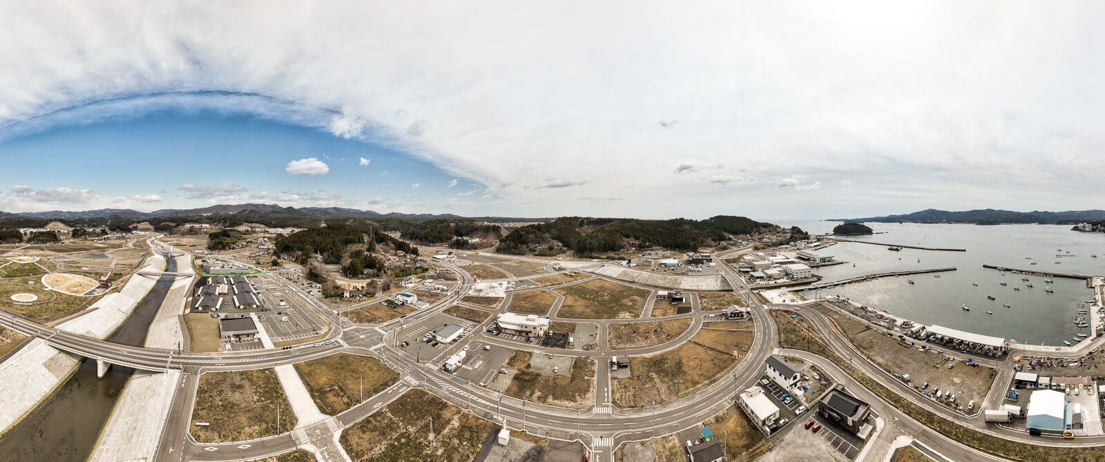 右側に広がる海と港、左側には整備された川と区画整備が進む土地が一体となって写されており、復興による都市の再構築と港町の風景が広がる港と街並み全体の昼間の風景を高台から定点カメラで撮影した写真