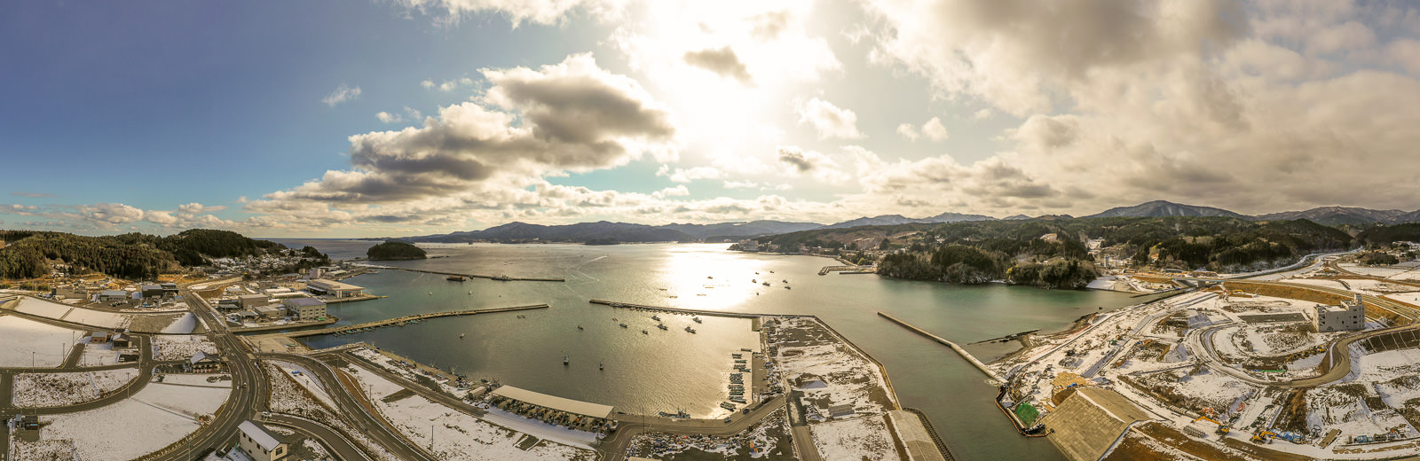 雪が積もった陸地に道路や建物が広がり、複数の桟橋が海に向かって伸び、港には船が停泊しており、背景には山々が連なり、雲の合間から日差しが差し込む様子が見える冬の海辺の港町を広く見渡した写真
