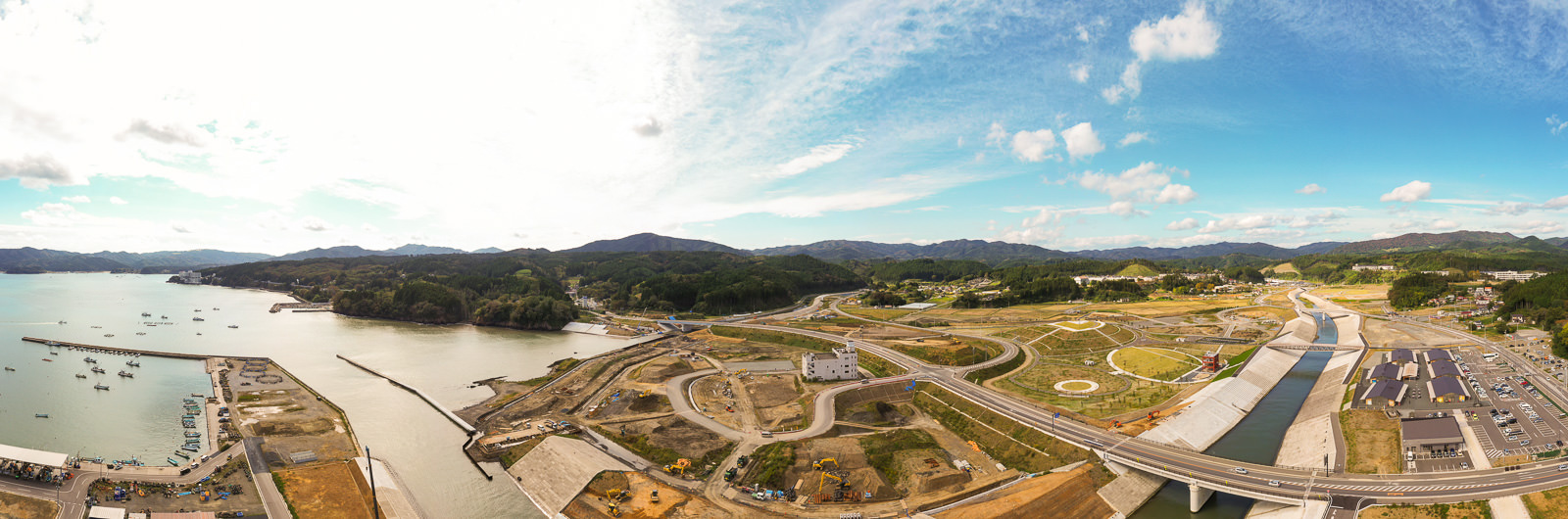 左側に海が広がり、複数の船が停泊する水辺に沿って、道路や橋、建設中または新しく建てられた建物が点在し、背景には丘や山々が広がる沿岸開発地域の写真