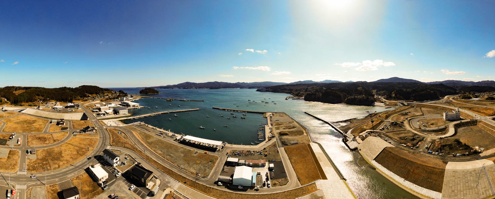 港を中心とした海岸地域のパノラマ風景が広がっており、水面には複数の船が浮かび、海岸線にはさまざまな建物やインフラが並び、道路や駐車場、建設現場も点在し、背景には丘や山々が広がり、空は澄んだ青空で太陽が明るく輝いている写真