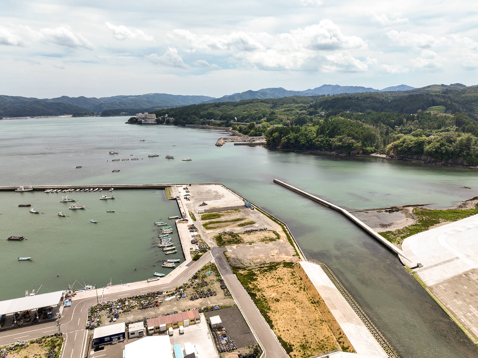 港湾の突堤と防波堤が湾を囲み、小型船舶が多数停泊し、背後に緑豊かな山々が広がる景観の昼間の風景を高台から定点カメラで撮影した写真