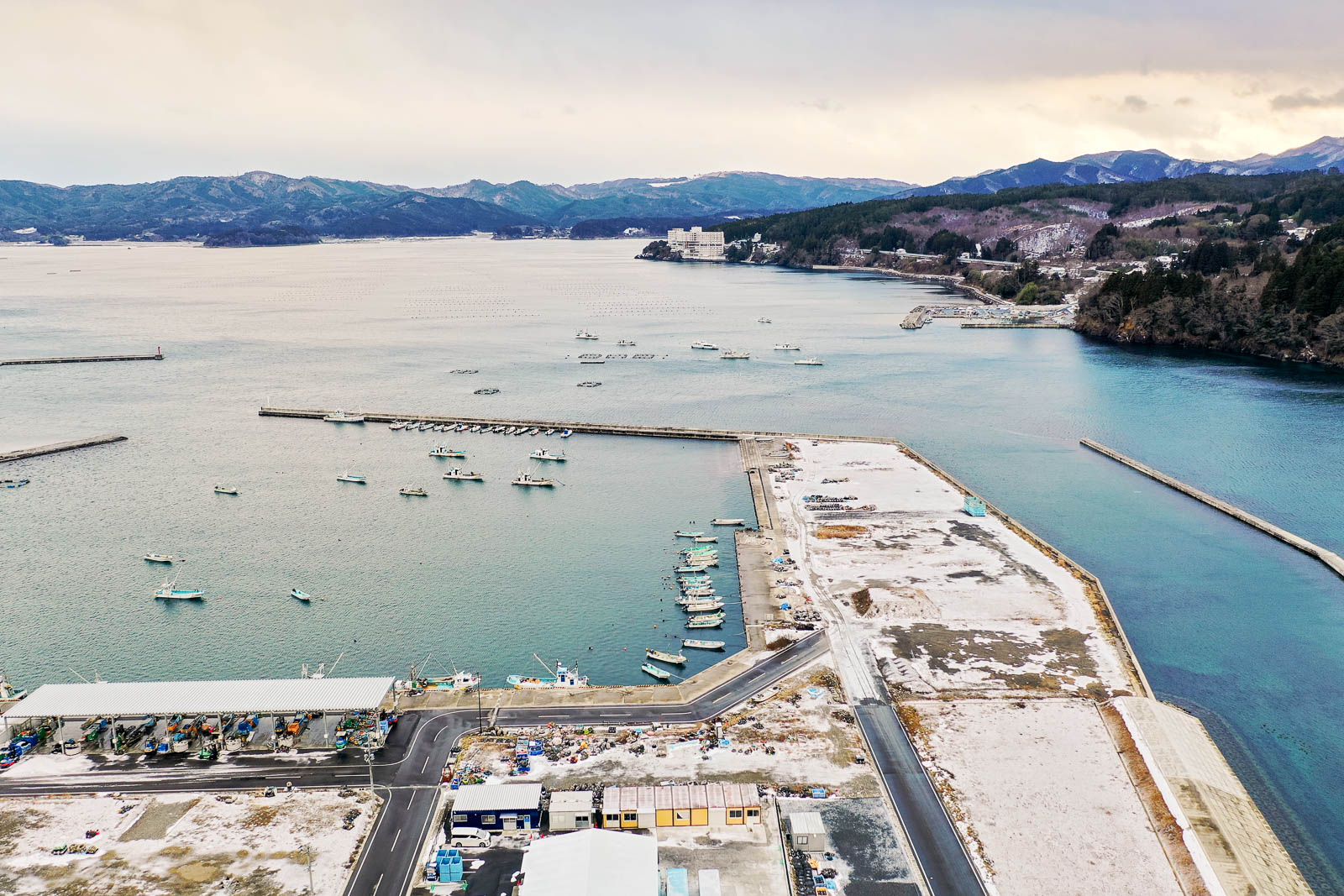 漁船が浮かぶ静かな港と岸壁、遠くにリゾートホテルのような白い建物と山が見える昼間の風景を高台から定点カメラで撮影した写真