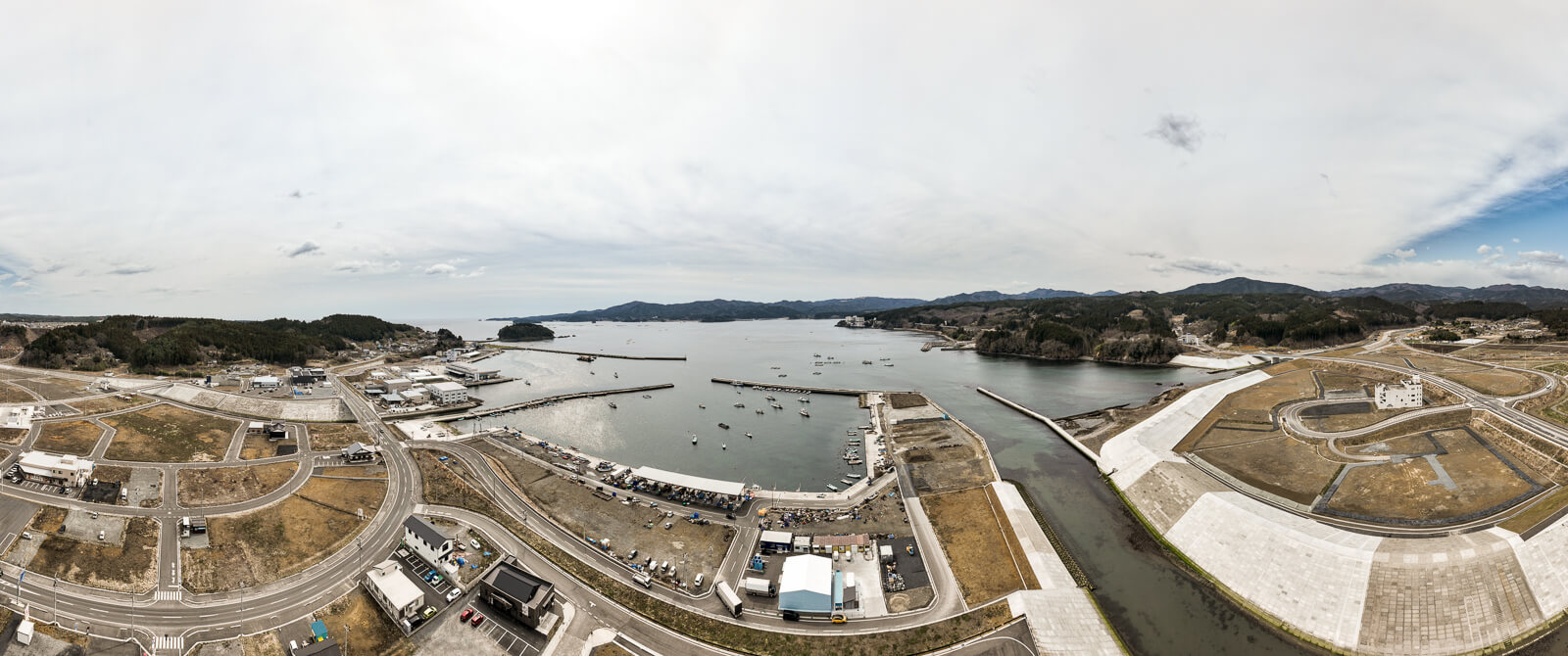 防波堤に囲まれた湾内には多くの船が浮かび、港周辺には倉庫や建物が建ち並ぶ、湾と漁港の昼間の風景を高台から定点カメラで撮影した写真