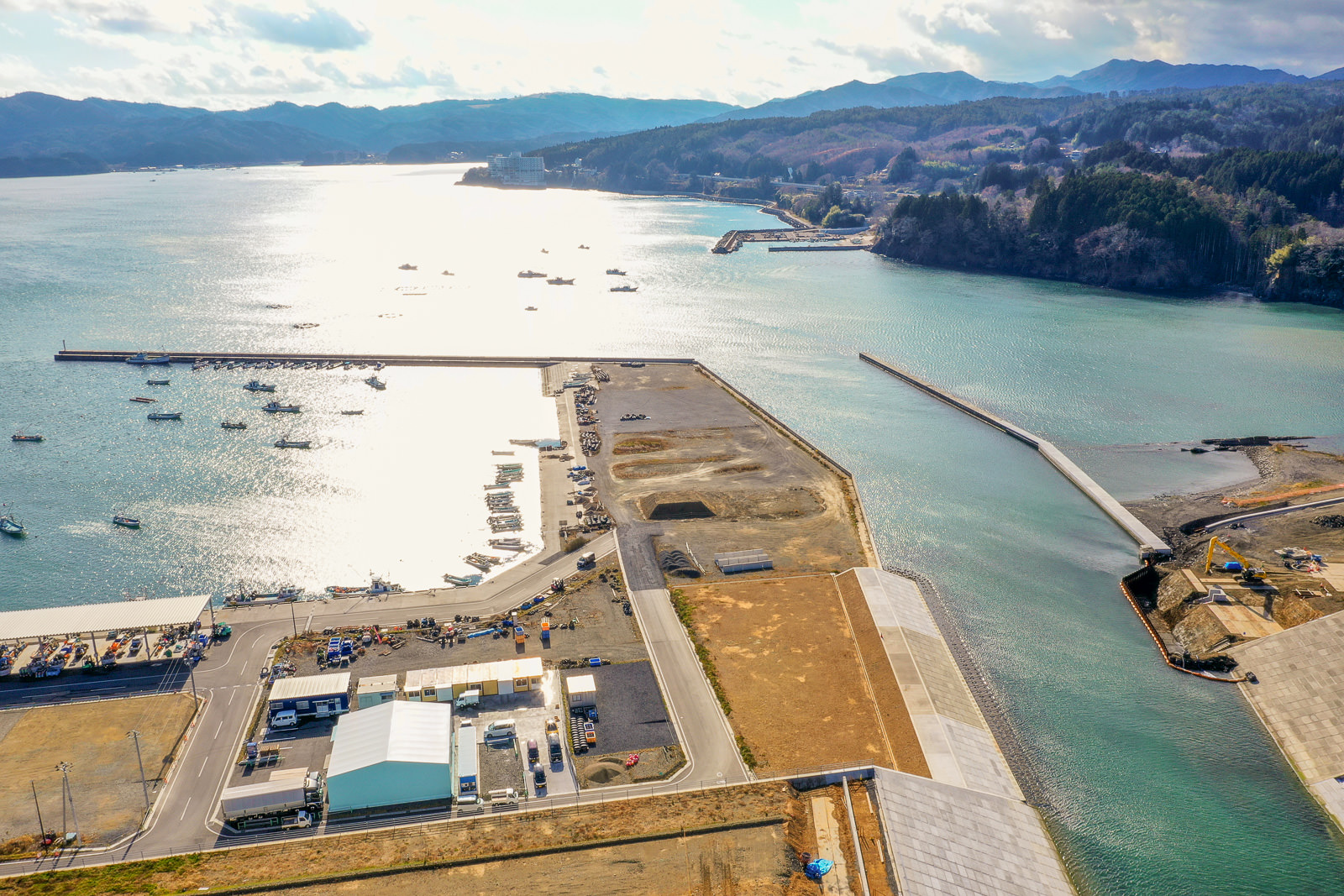 三方を陸地に囲まれた港を中心とした海岸地域を上空から撮影した風景で、海には複数の船が停泊しており、陸地には建物や道路、建設中の場所が点在し、水面は太陽光を反射して輝き、遠くには山々が広がる港湾地域の写真