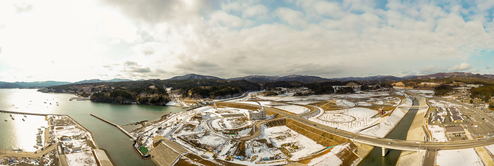 雪が積もった海沿いの地域を広く見渡す構図で、曲がりくねった道路や橋、水路が入り組み、背景には丘や山が連なり、空には雲が点在する冬の海辺の風景の写真