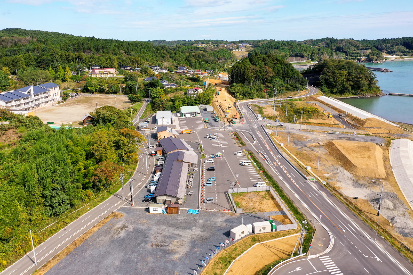 海岸沿いに広がる地域を空から捉えた風景で、数軒の建物、駐車場と車両、小規模な建物が点在し、片側には密集した森林、もう一方には海岸が広がり、沿岸部では建設作業が行われ、背景には丘陵が見える沿岸開発地域の写真