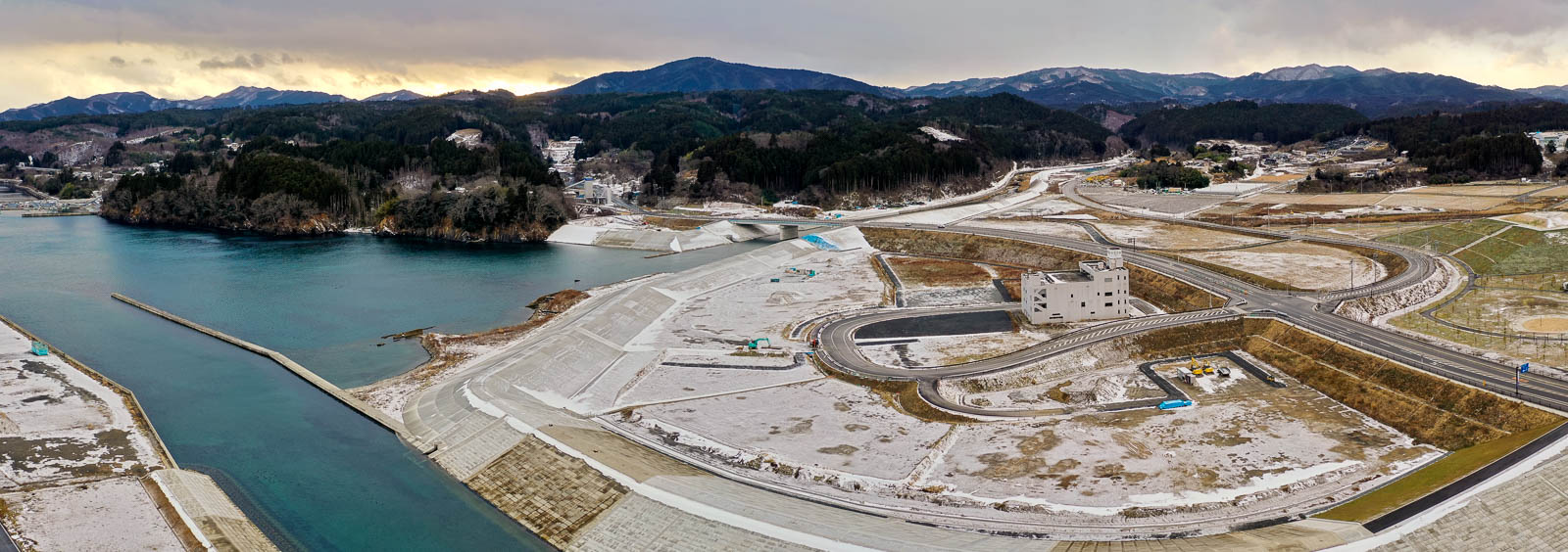 海に面した湾の入り口に防波堤が伸び、周囲を山々と森が囲み、雪の積もる護岸と河川改修工事の様子が広がっている昼間の風景を高台から定点カメラで撮影した写真