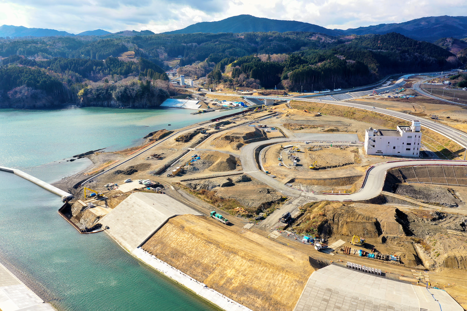 水辺の近くに広がる大規模な建設現場を上空から撮影した風景で、敷地内には曲がりくねった道路や小道が走り、建設機材や車両が点在し、背景には森林に覆われた丘や山が広がり、右側には白い建物が見え、空には雲が浮かぶ建設現場の写真