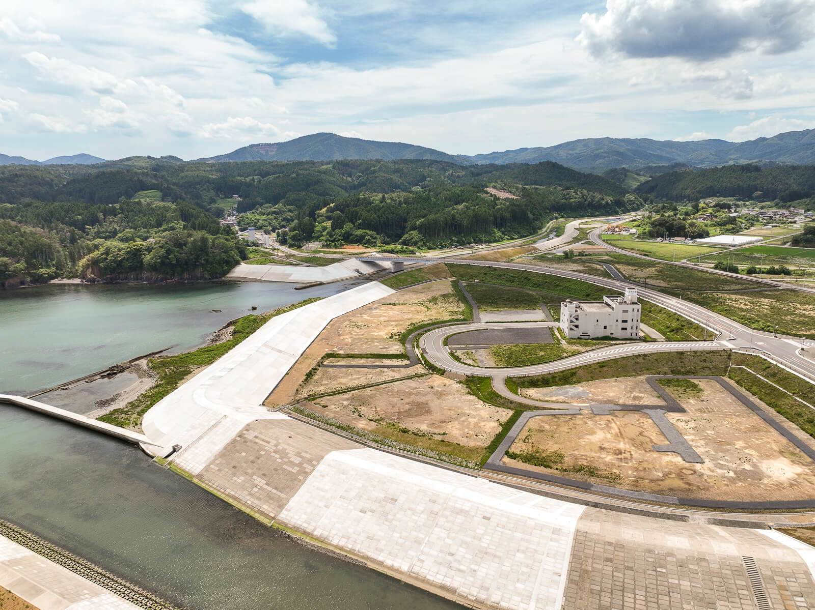 白く舗装された川の堤防が湾岸まで続き、中央に立つ白い建物の周囲は整地された土地と道路、背後に山々が見える昼間の風景を高台から定点カメラで撮影した写真
