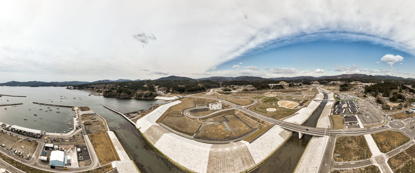 湾と川が交わる地点から見た全体風景で、左右に防波堤と護岸が延び、中央に川がまっすぐ流れ、両側には都市再開発された土地と緑地、道路が整然と配置されている復興都市全体の昼間の風景を高台から定点カメラで撮影した写真