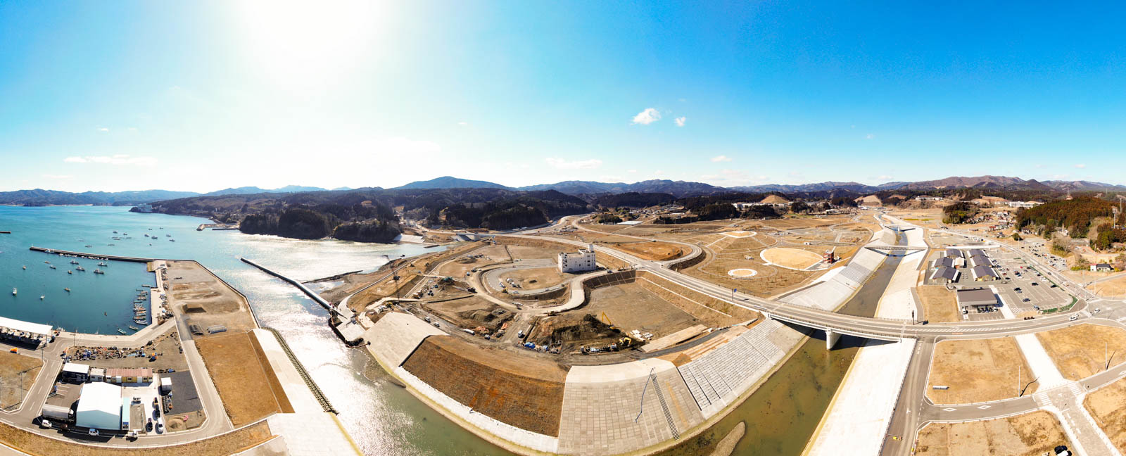 海岸地域を上空から見下ろした風景が広がっており、左側には水域が広がり、複数の船が桟橋に停泊していて、陸地には道路や橋、堤防などの治水施設が整備され、建物や広場も点在し、背景には丘や山々が広がり、空は澄んだ青空が広がっている写真
