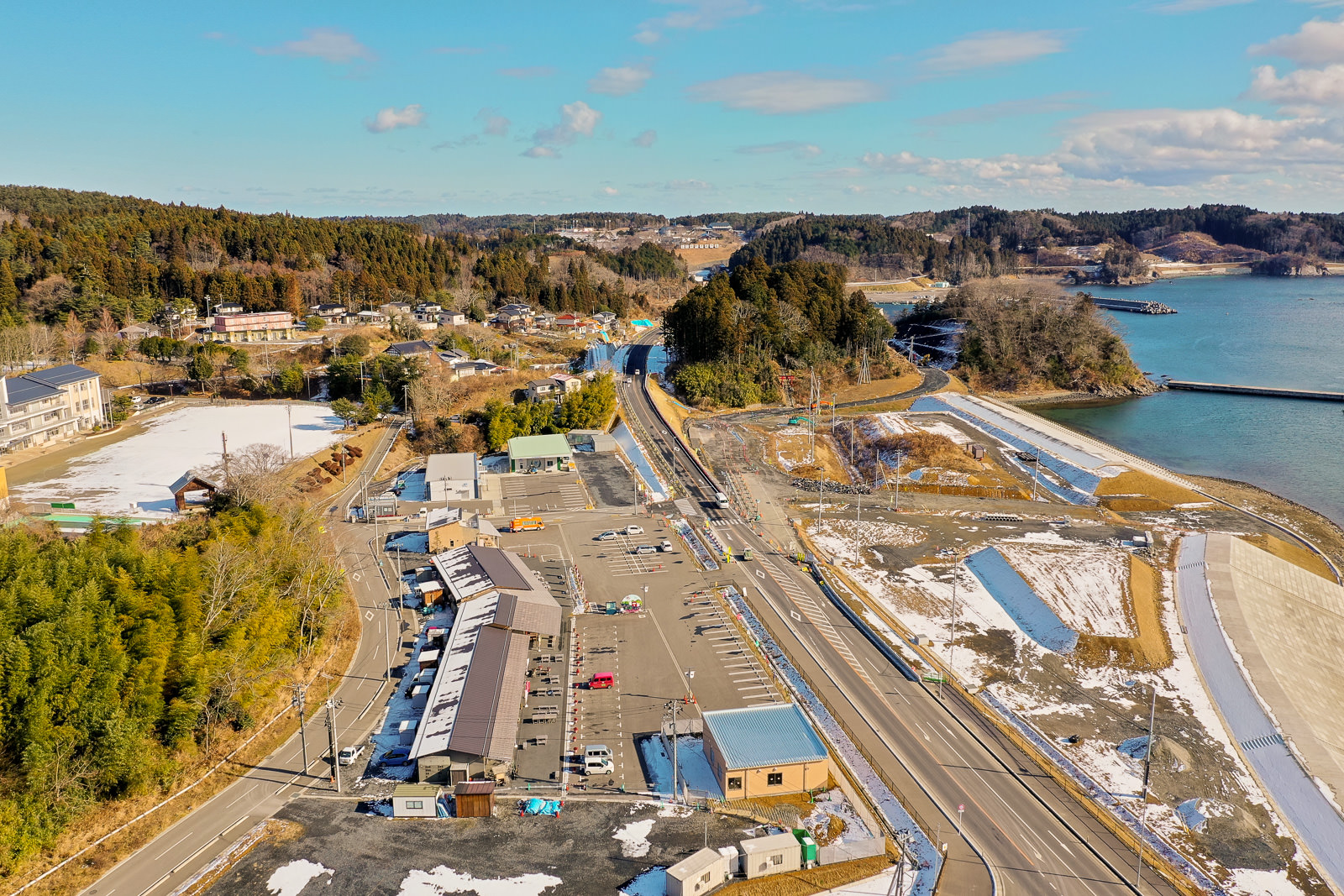 建物や道路、自然の風景が入り混じる海沿いの地域を上空から撮影したもので、複数の構造物の中には駐車場があり、車が停まっていて、周囲には木々や丘が広がり、右側には海岸線とその先に広がる水面が見え、地面には雪がまだらに積もり、空は雲が少し浮かぶ晴れた冬の日の海辺の町の写真