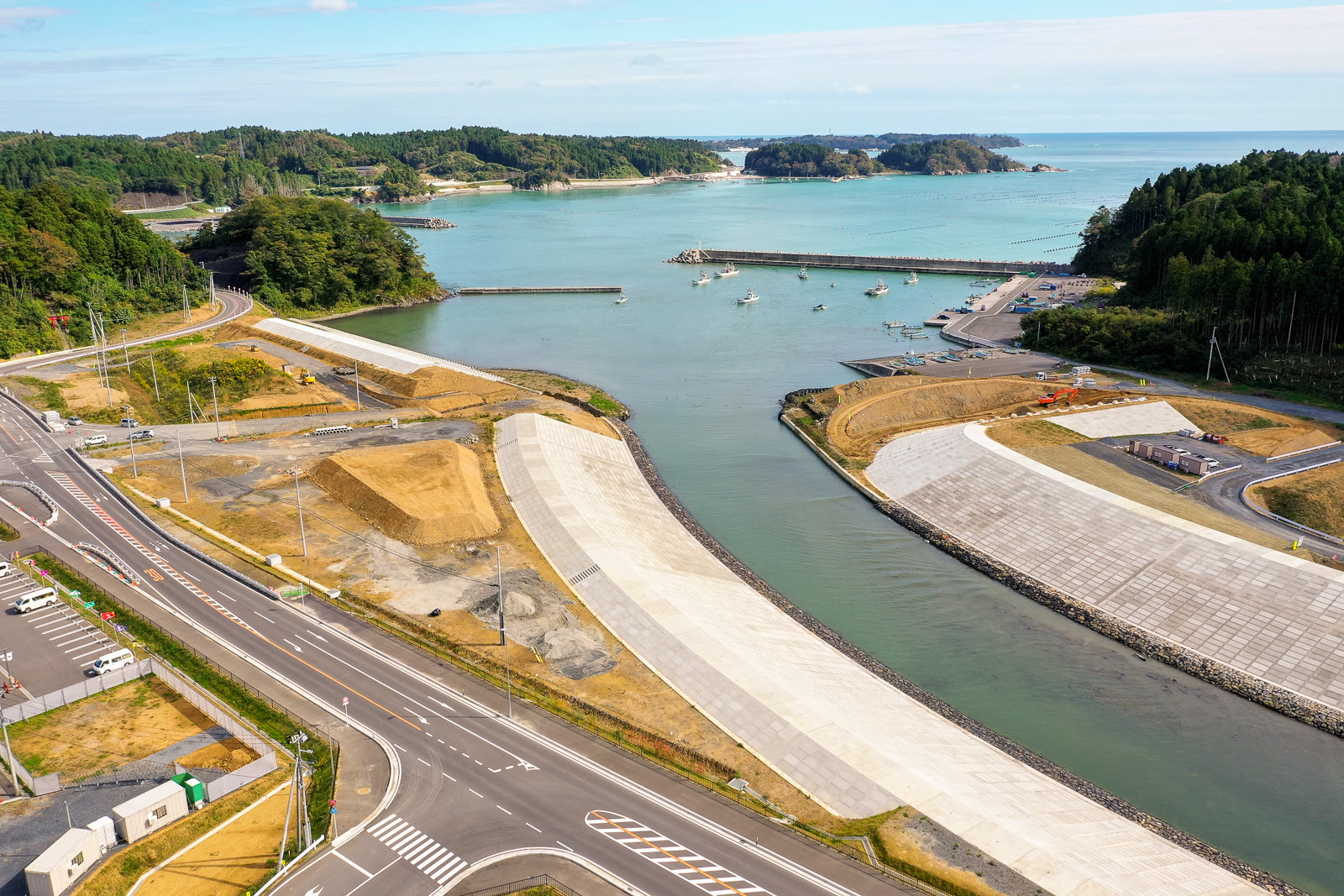 川が海へと流れ込む沿岸地域を空から捉えた風景で、道路や駐車場、建設現場が点在し、岸辺には複数の船が停泊し、周囲には緑豊かな森林と小さな丘が広がる海辺の地域の写真