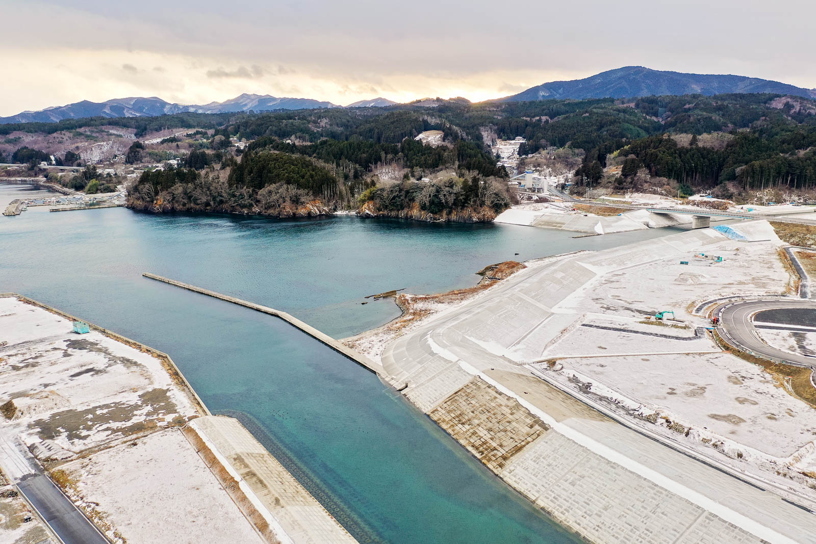 左側に海が広がり、湾の奥に森と山々が見え、手前にはコンクリートで整備された護岸と海岸線が続いている冬の港湾整備地域の昼間の風景を高台から定点カメラで撮影した写真